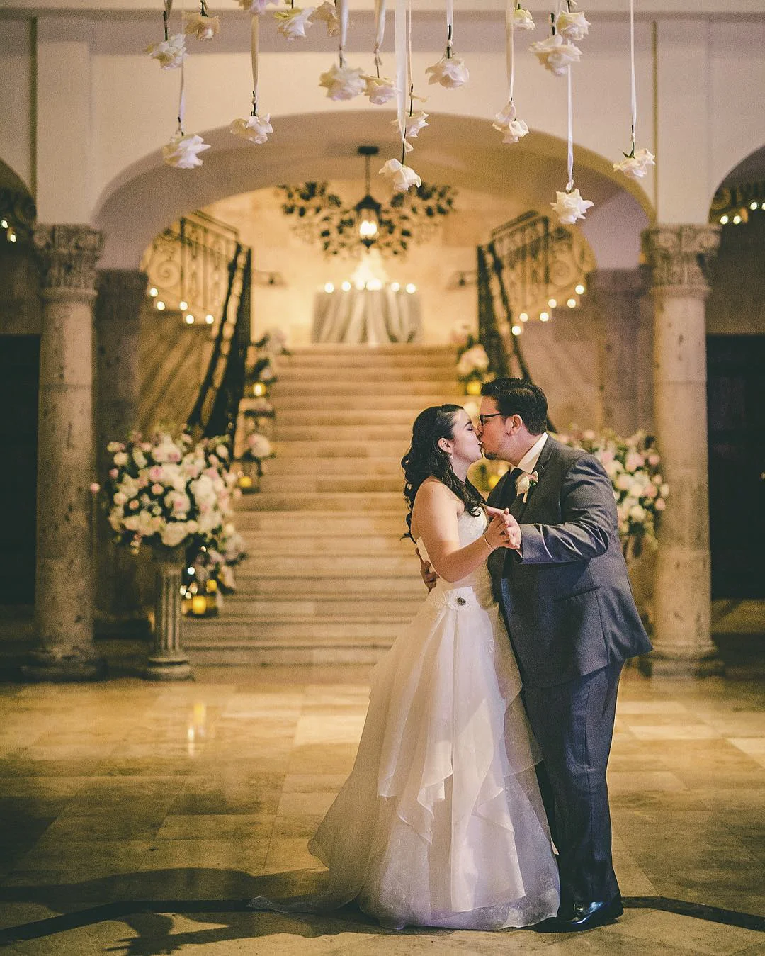 Newlywed couple dancing and kissing beneath hanging flowers with a grand staircase in the background.
