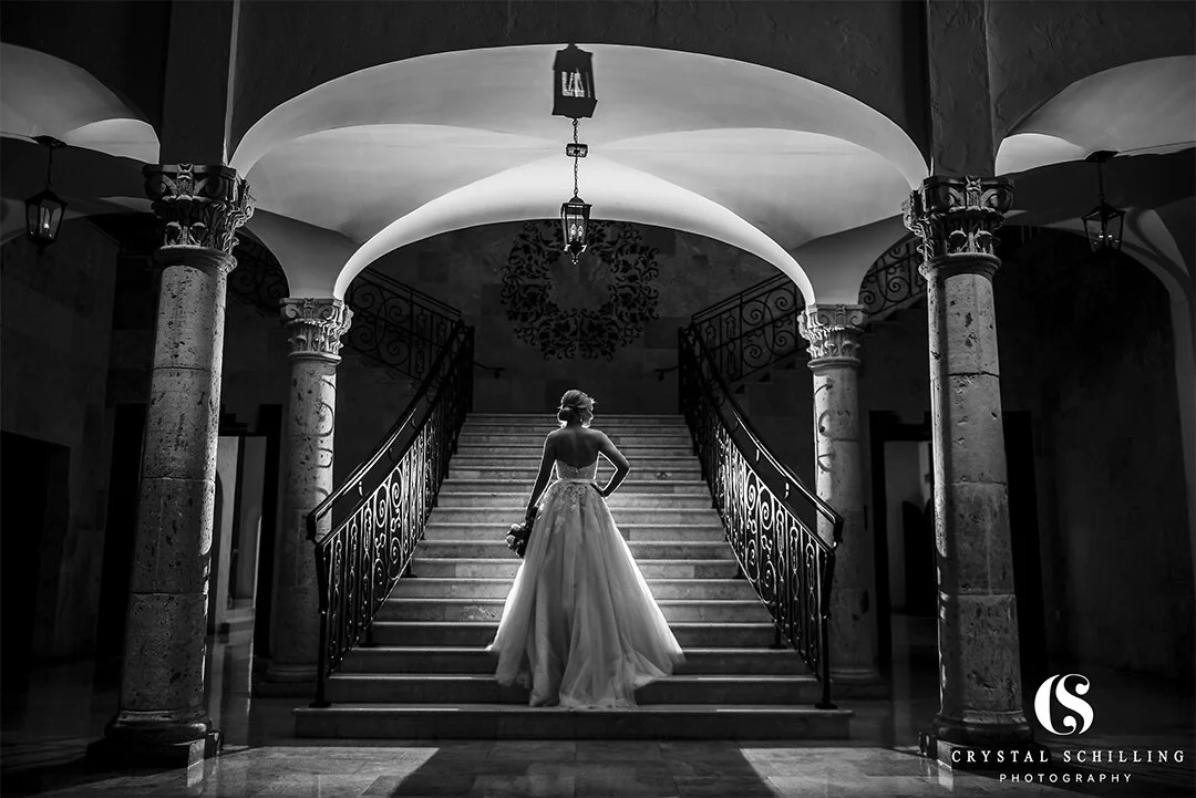 Black and white photo of a bride holding flowers, standing at the base of a grand staircase under ornate arches.