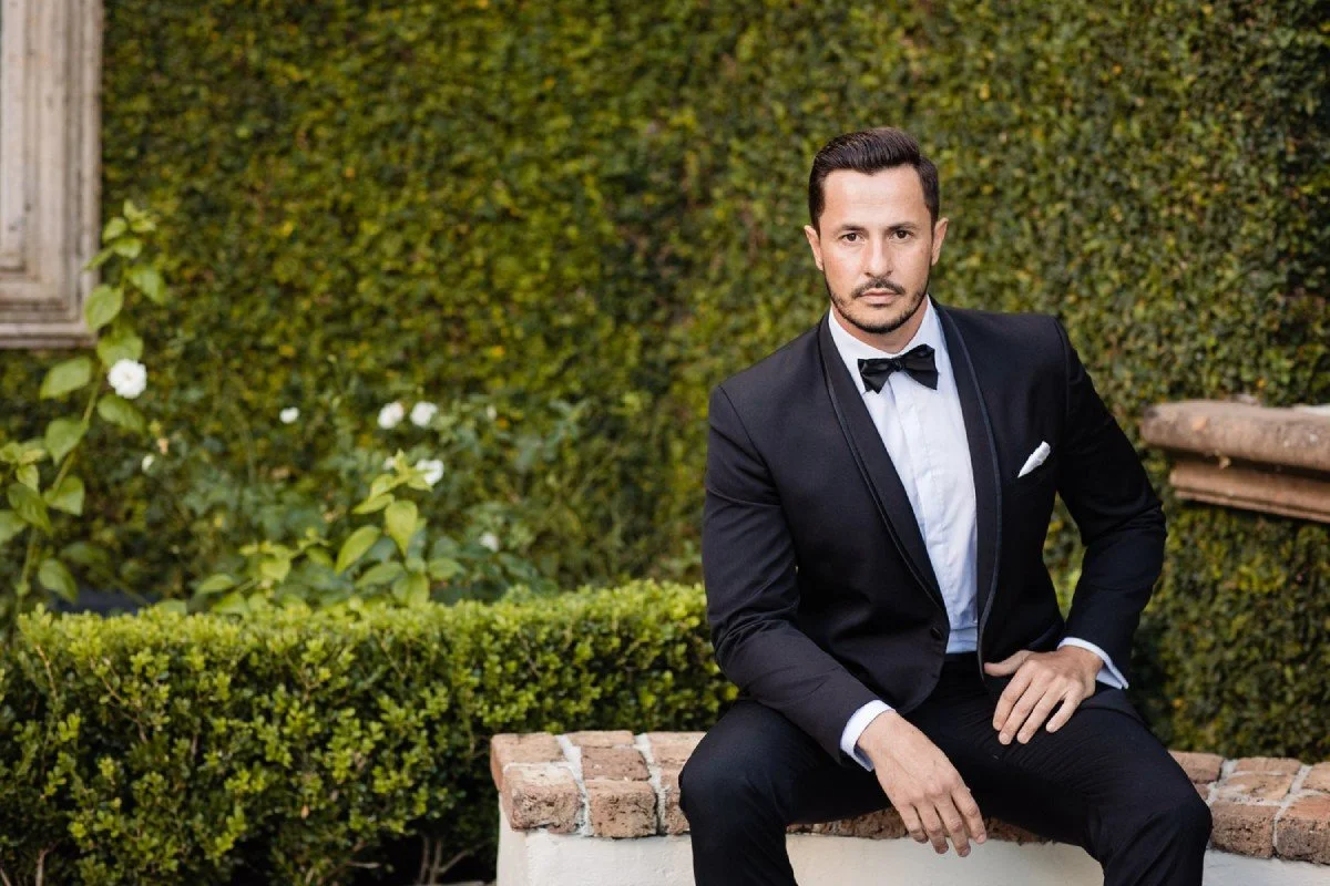 Man in black tuxedo with bow tie sitting on a garden bench, posing in front of lush greenery and white flowers.